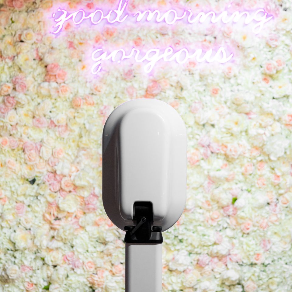 Modern digital photo booth setup with a floral wall backdrop and neon sign at an event