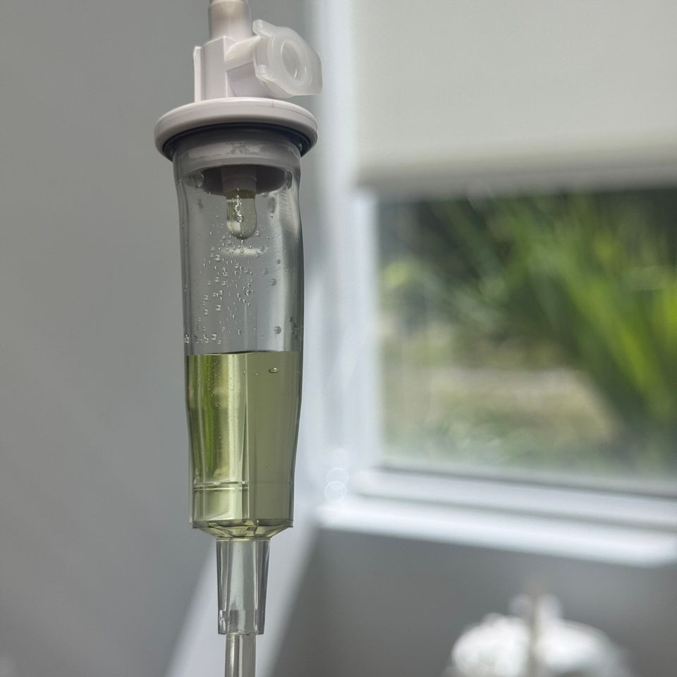 Close up of an IV drip chamber with yellow vitamin fluid beside a bright window