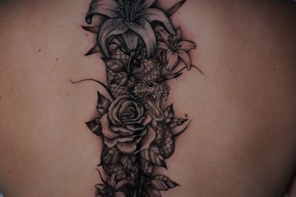 Image of a tattoo on a womans back that has a serpent ready to strike and hidden by the cover of very detailed rose and tulip in black and grey ink