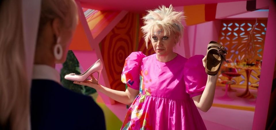 Kate McKinnon as "weird Barbie" with a pink pump in one hand and a Birkenstock in the other 