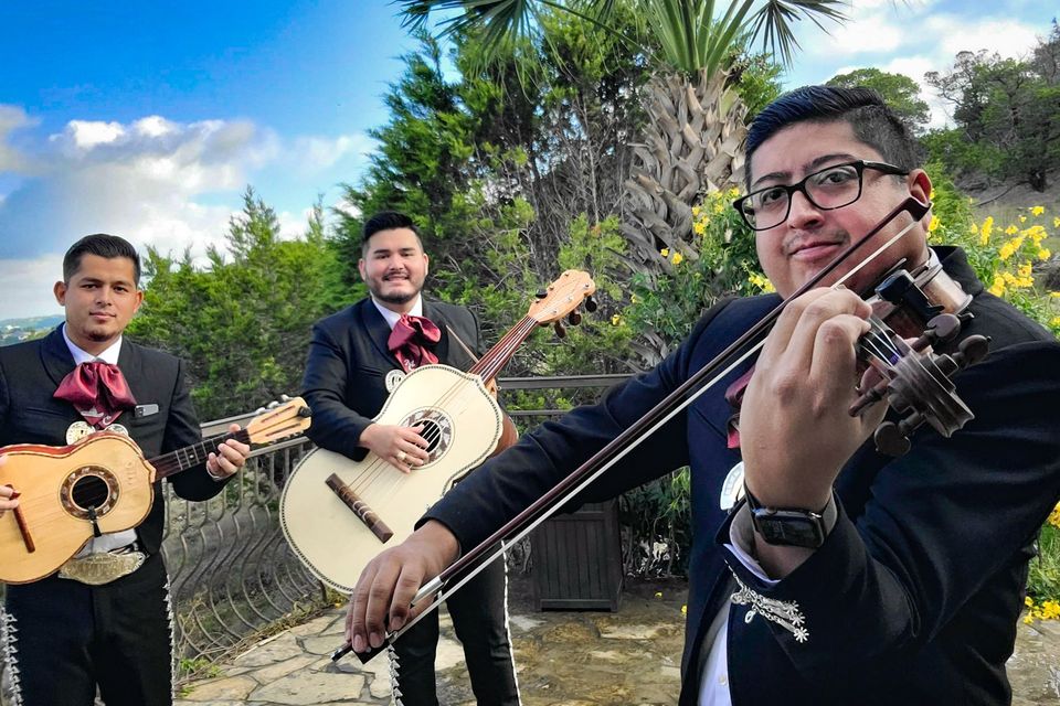 Three Mariachi Capital musicians in traditional black charro suits performing outdoors with violin, guitarrón, and vihuela.