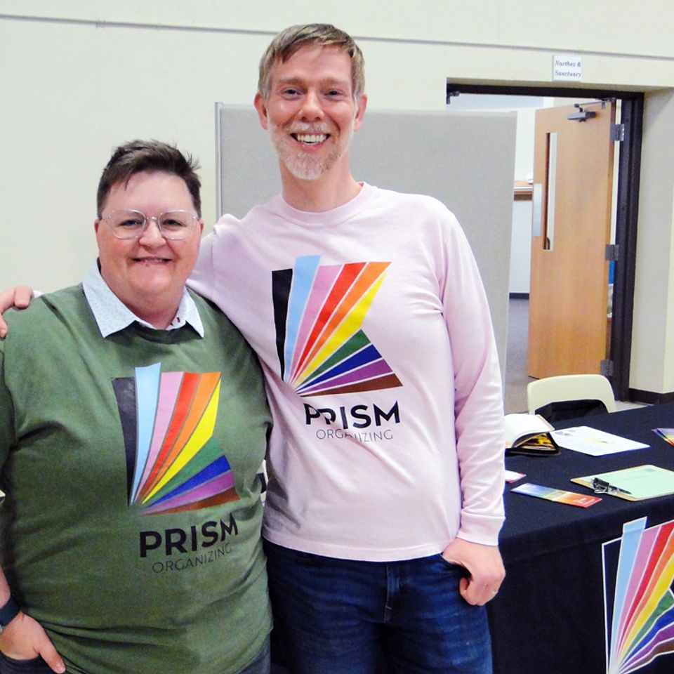 Two adults wearing Prism Organizing shirts in different colors in front of Prism Organizing booth