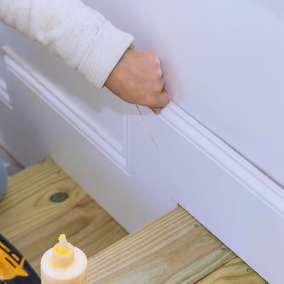Hand applying adhesive along white baseboard molding during a trim installation
