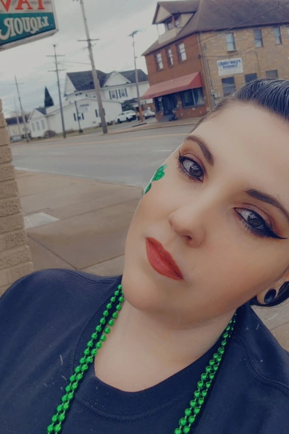 Young woman wearing green beads and shamrock face paint posing for a St. Patrick’s Day selfie.